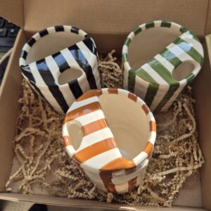 A close-up photo of a handmade ceramic mug with a smooth, gloss glaze in soft off-white, sitting in a Brown kraft box. The mug has a rounded body, a sip cup detailing and it is handleless. The natural light casting gentle shadows that highlight its colourway of Black, Green or Brown.