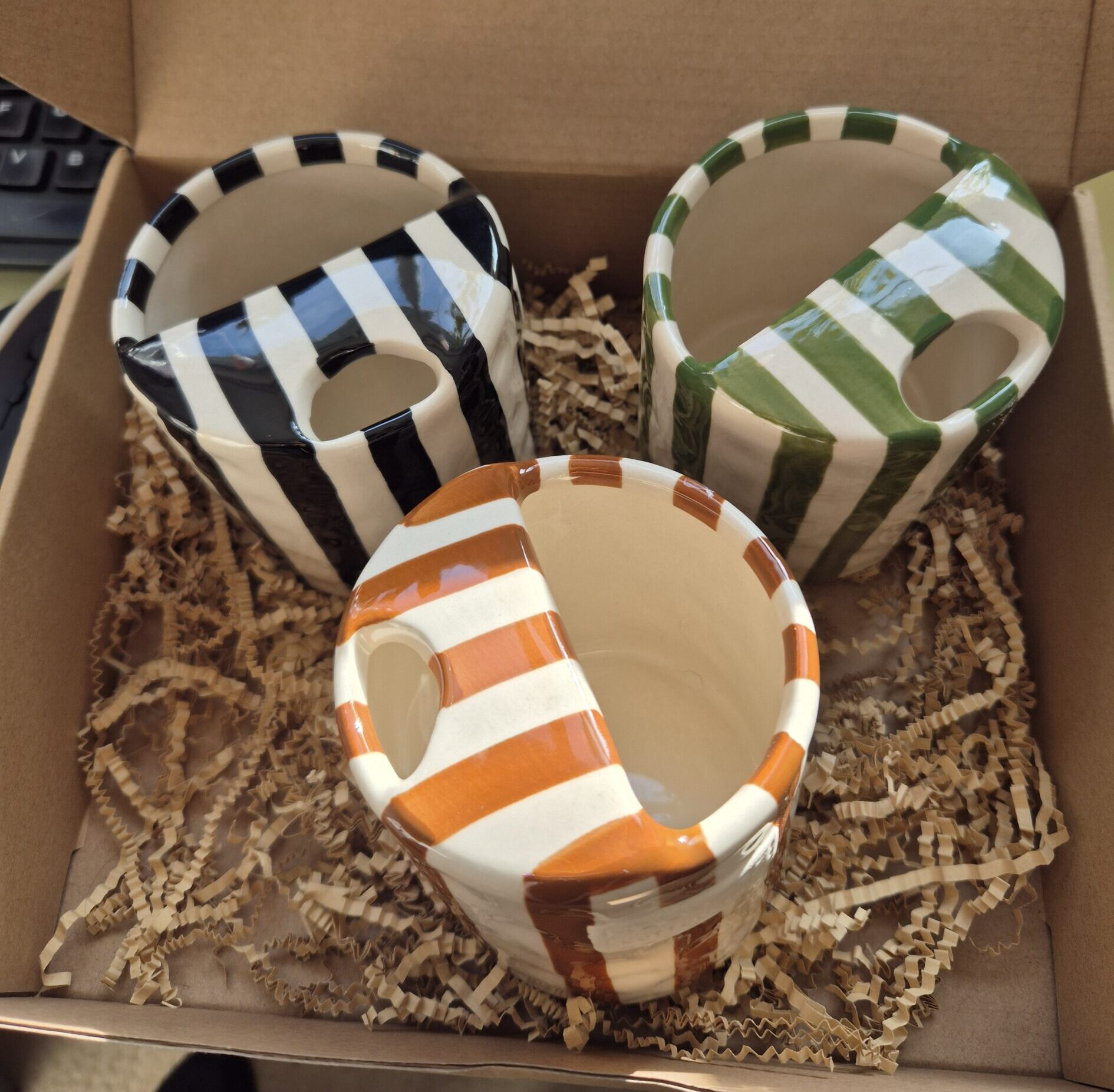 A close-up photo of a handmade ceramic mug with a smooth, gloss glaze in soft off-white, sitting in a Brown kraft box. The mug has a rounded body, a sip cup detailing and it is handleless. The natural light casting gentle shadows that highlight its colourway of Black, Green or Brown.