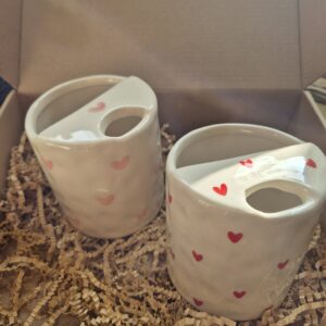 A close-up photo of a handmade ceramic mug with a smooth, gloss glaze in soft off-white, sitting in a Brown kraft box. The mug has a rounded body, a sip cup detailing and it is handleless. The natural light casting gentle shadows that highlight its colourway of Red love hearts or Pink love hearts.