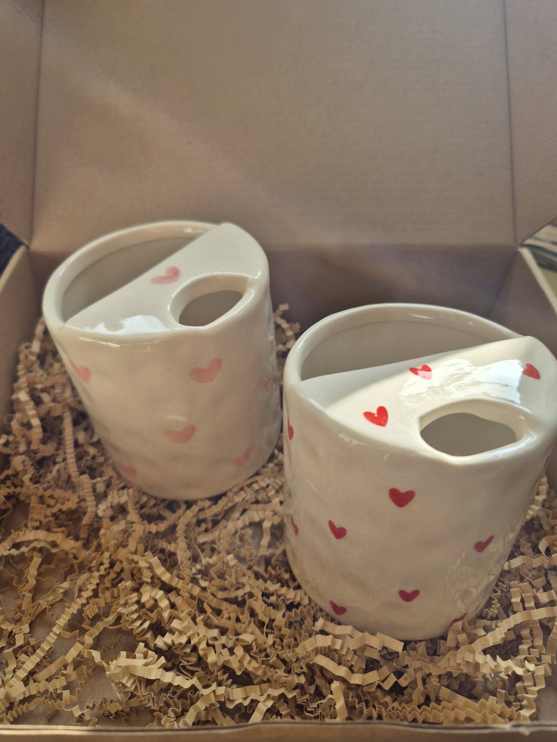 A close-up photo of a handmade ceramic mug with a smooth, gloss glaze in soft off-white, sitting in a Brown kraft box. The mug has a rounded body, a sip cup detailing and it is handleless. The natural light casting gentle shadows that highlight its colourway of Red love hearts or Pink love hearts.