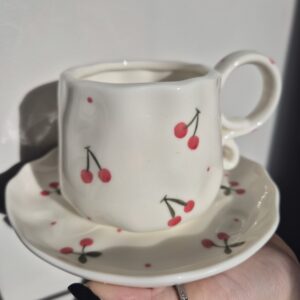 A close-up photo of a handmade ceramic mug with a smooth, gloss glaze in soft off-white. The mug has a rounded body, with a unique spiral handle. The natural light casting gentle shadows that highlight the Red cherries all over the mug