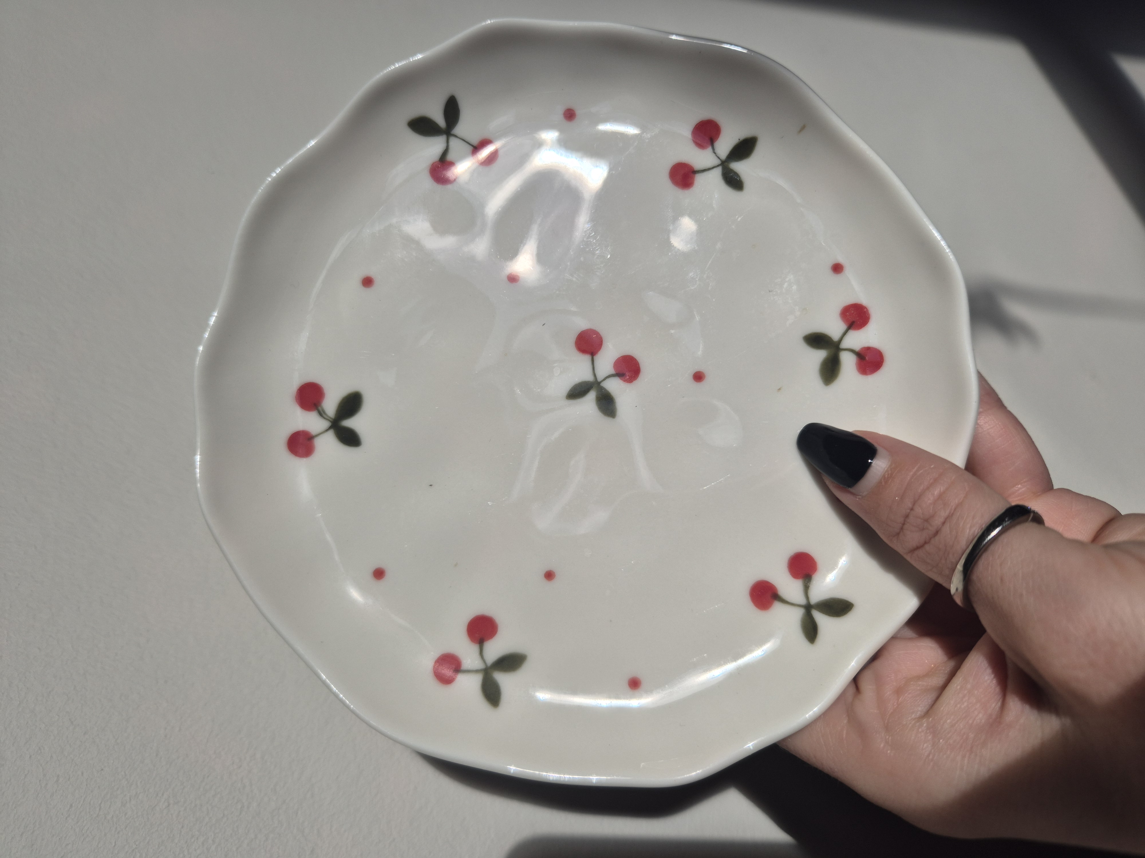A close-up photo of a handmade ceramic side plate with a smooth, gloss glaze in soft off-white. The plate has a rounded body with small Red and Green cherries and stalks all over