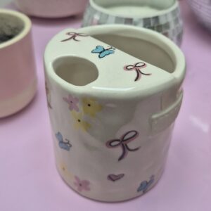 A close-up photo of a handmade ceramic mug with a smooth, glossed glaze in soft off-white, sitting on a Pink background. The mug has a rounded body, a sip cup detailing and it is handleless. The natural light casting gentle shadows that highlight its handpainted detailing of Bows, Butterflys, hearts and flowers.