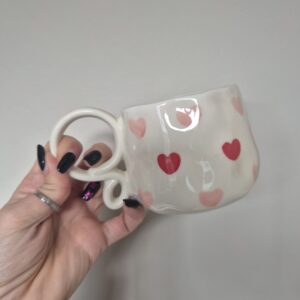 A close-up photo of a handmade ceramic mug with a smooth, gloss glaze in soft off-white. The mug has a rounded body, with a unique spiral handle. The natural light casting gentle shadows that highlight the Pink and Red love heart detailing.