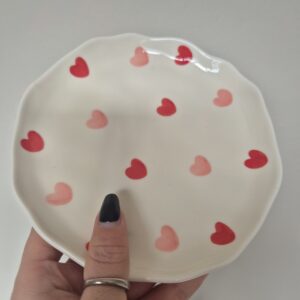 A close-up photo of a handmade ceramic side plate with a smooth, gloss glaze in soft off-white. The plate has a rounded body with small Red and Pink hearts all over